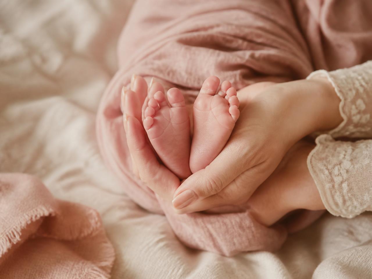 A parent's hands cradling a newborn baby's feet on soft pink linen